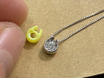 石留め直前のプラチナ製馬蹄形ペンダントと、その横に置かれた黄色い3Dプリント樹脂の原型。ベネチアンチェーンが通っている。