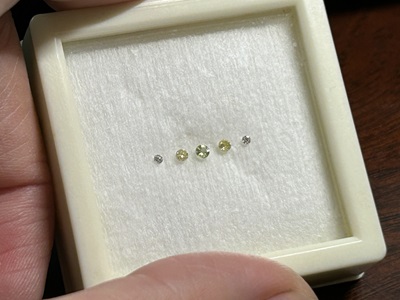 作業マットの上にピンセットで整列された、色の異なる5石の小さなルース。隣には金属の指輪パーツ。