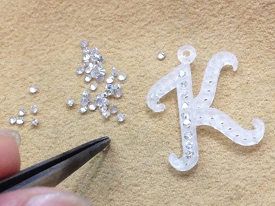 「K」の樹脂原型と、その横にピンセットで並べられた本物のメレダイヤモンド。