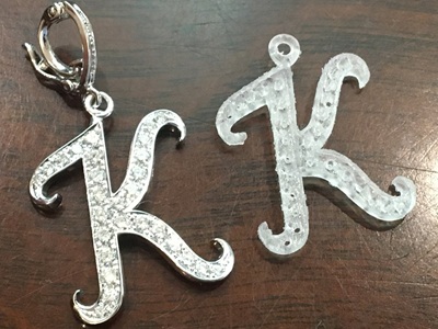 完成したプラチナ製の「K」ペンダントと、樹脂原型が並んでいる様子。ペンダントには上部にオープンバチカンが付いている。