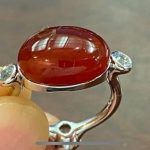 完成したレッドアンバーリング。光にかざされ、半透明のアンバーの奥から、設計された蝶のシルエットがふわりと浮かび上がっている様子。両サイドのダイヤもきらめく。