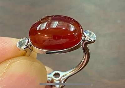 完成したレッドアンバーリング。光にかざされ、半透明のアンバーの奥から、設計された蝶のシルエットがふわりと浮かび上がっている様子。両サイドのダイヤもきらめく。