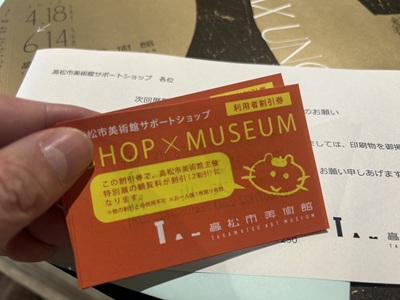 手に持ったオレンジ色の「高松市美術館サポートショップ 利用者割引券」のカード。