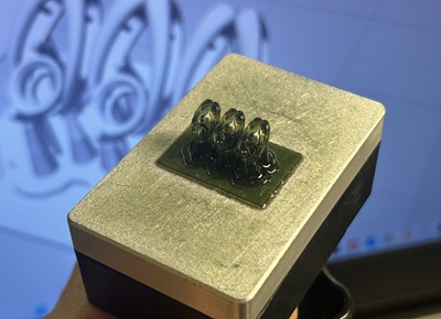 3Dプリンターのプラットフォーム上に、緑色の透明なレジン樹脂で細部まで精密に出力された3つのペンダント原型の写真