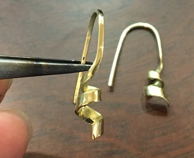 ピンセットで挟まれた、鋳造から上がって美しく磨きをかけられたリボン型のK18金製（750）フックピアスのパーツ