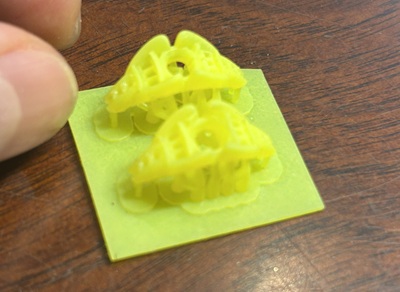 木目のテーブルの上に置かれた、黄色の樹脂製3Dプリントパーツ。リングの上部(石座部分)の形状をした試作品が2点並んでいる