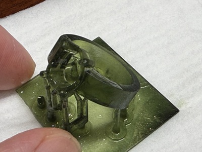 3Dプリントされた指輪の原型のクローズアップ。人差し指と親指で支えられており、複雑な石座のデザインや、造形を支える細いサポート材が確認できる。