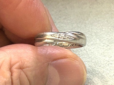 指先で持った結婚指輪のアップ。中央に3石のメレダイヤがあり、内側には記念日などの文字が刻印されている。