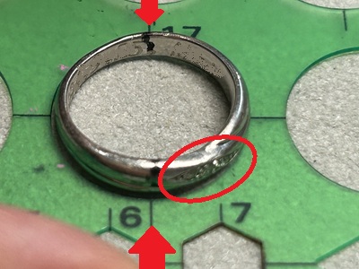 緑色のテンプレート定規の上に置かれた結婚指輪。切断予定の2箇所に黒いマークが施され、赤い円と矢印で位置が強調されている。