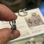 指先で持った完成済みのプラチナメレダイヤピアスと、その背景に写るラミネート加工されたカード型のリメイク証明書。