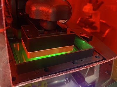 3Dプリンターの内部写真。赤いアクリルカバー越しに、緑色の液体レジンが満たされたトレイへ造形プラットフォームが浸かり、底面から緑色のUVレーザー光が照射されて原型が精密に硬化・造形されている様子。