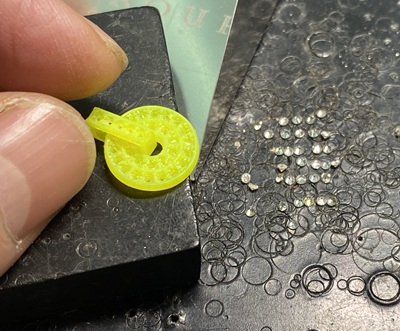 職人の指先で持たれた、3Dプリンターから出力された鮮やかな蛍光黄色の樹脂製サークルペンダント原型。右側の黒い作業台の上には、セットされる予定の小さなメレダイヤモンドが多数並べられている状態の写真。