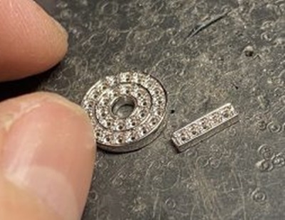 黒い作業マットの上に置かれた、金属へと鋳造されたプラチナ(Pt)製のサークルパーツとバチカン(縦長のパーツ)。ダイヤモンドを留めるための小さな穴や爪の土台が綺麗にキャストされている状態の写真。