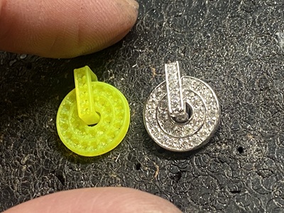 黒い作業マットの上に並べられた、蛍光黄色の樹脂製ペンダントパーツと、すべての石留めを終えてまばゆく輝くプラチナ製のペンダントパーツの比較画像。スクエアのバチカン部分にも細かくダイヤモンドが敷き詰められている状態。
