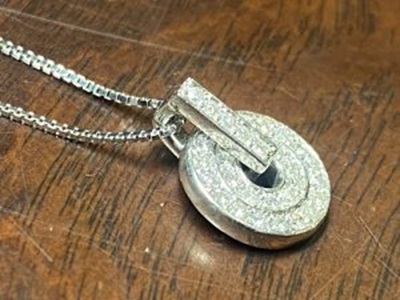 木目の美しいテーブルの上に斜めに置かれた、完成した立体円盤型のダイヤモンドペンダントトップ。ベネチアンチェーンが繋がり、プラチナの立体的な厚みとパヴェダイヤモンドの眩い煌めきが絶妙に調和している斜めからの完成写真。