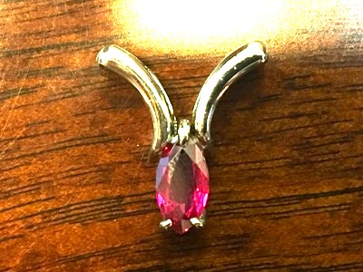 木目調のテーブルの上に置かれた、完成したプラチナ製のペンダントトップ。美しい赤紫色のマーキスシェイプルビーがしっかりと留められ、イニシャル「Y」をモチーフにしたアーム部分が上品に輝いている実物写真。