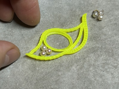 作業マットの上で、3Dプリンターから出力された蛍光黄色の樹脂製ブローチ枠を指で支えながら、中央の枠の左側に3粒のベビー真珠を乗せてサイズ感を確かめている手元の写真。