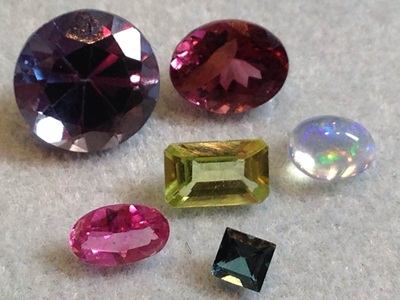 グレーのマットの上に並べられた6石の美しい宝石のルース。大粒の紫がかった青い石、赤紫のオーバル石、遊色効果のある乳白色のオパール、長方形のエメラルドカットが施された黄緑色の石、鮮やかなピンクのオーバル石、そして小さなスクエアカットの深い青緑色の石が写っている写真。