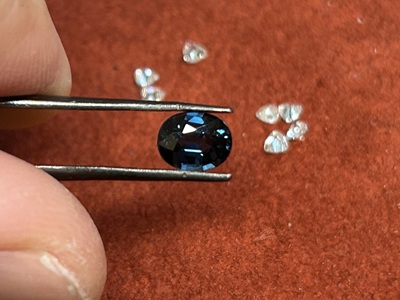 ピンセットでつまみ上げられた深みのある美しい濃紺のオーバルカットサファイア。背景には輝くメレダイヤモンドが数石置かれている写真。