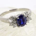 クリスタルのような透明な台の上に乗せられた、完成したプラチナ製のサファイアリング。中央の深みのあるサファイアを、両脇のダイヤモンドの葉っぱたちが華やかに引き立てている実物写真。