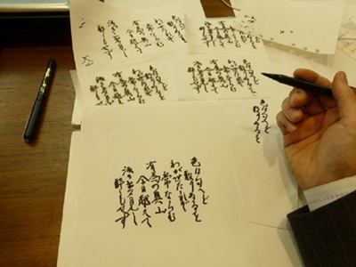 筆で「いろは歌」の清書を執筆している手元とデスクに並べられた書道用紙