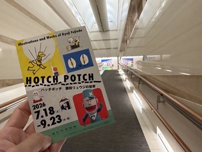 高松市美術館のロングスロープで特別展「ハッチポッチ 藤枝リュウジの世界」のチラシを手にする様子